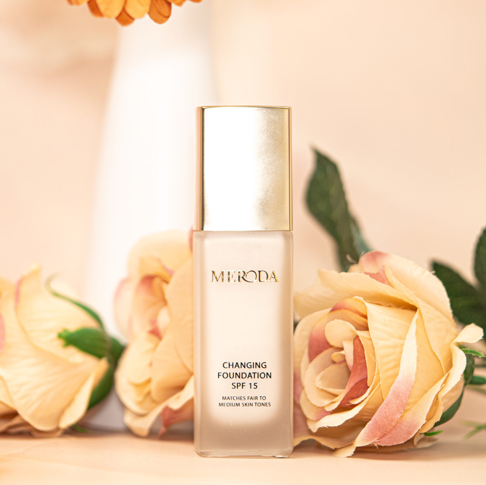 A bottle of Meroda Changing Foundation sitting on a peachy nude background with similar coloured flowers. 